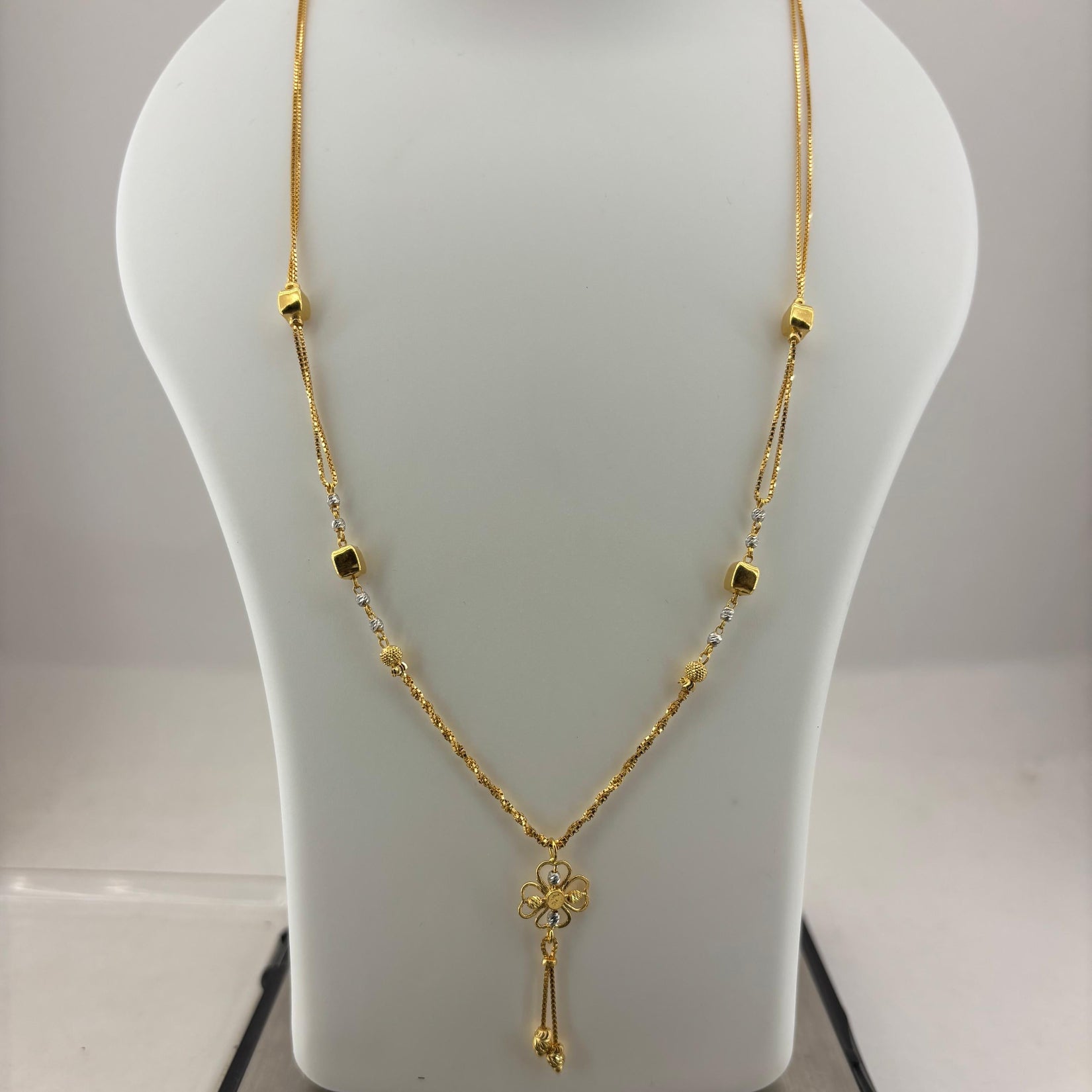 A gold necklace with a small floral pendant and dangling tassel, displayed on a white mannequin bust with a digital scale underneath.
