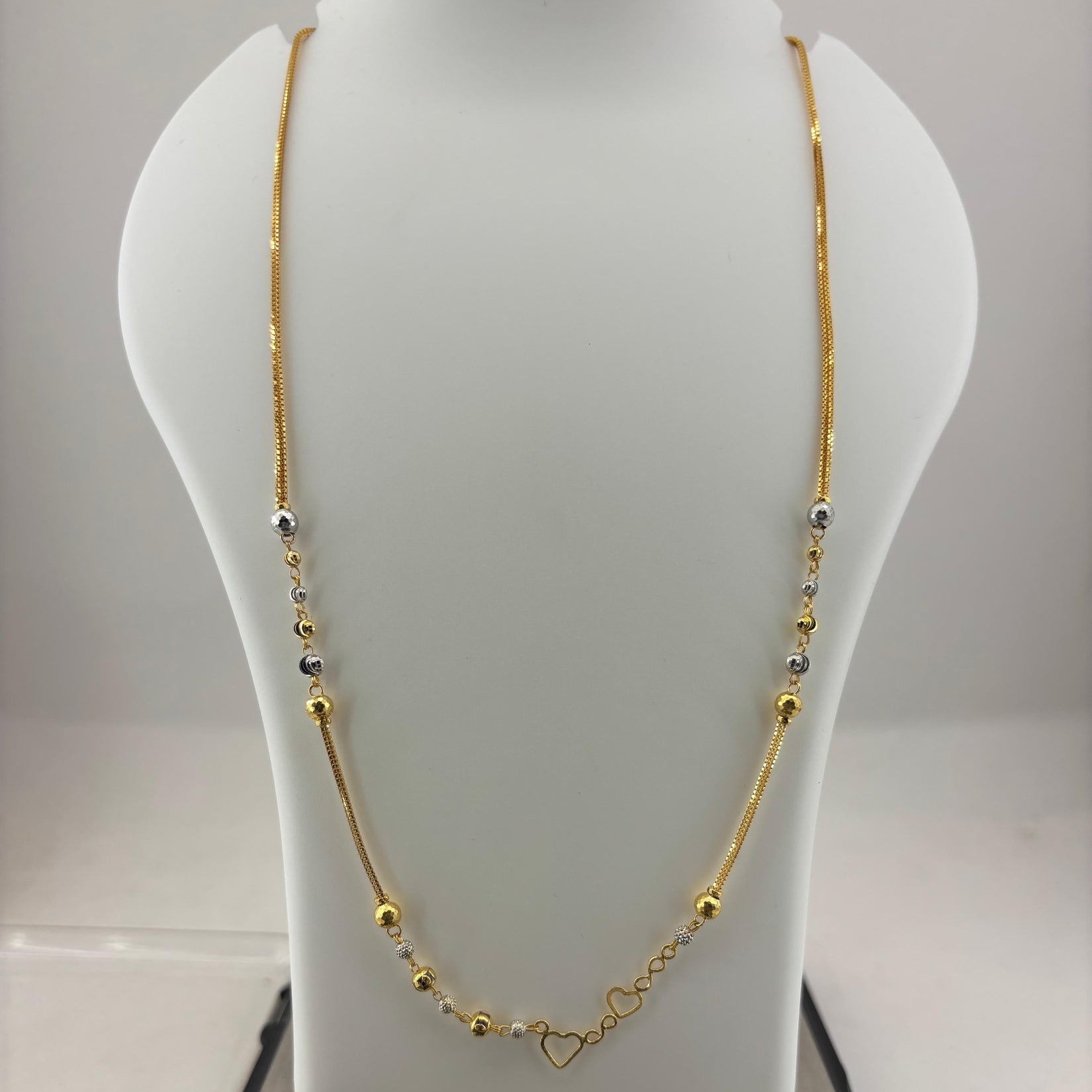 A gold necklace with small silver and gold beads and a heart-shaped clasp, displayed on a white jewelry bust sitting on a digital scale.