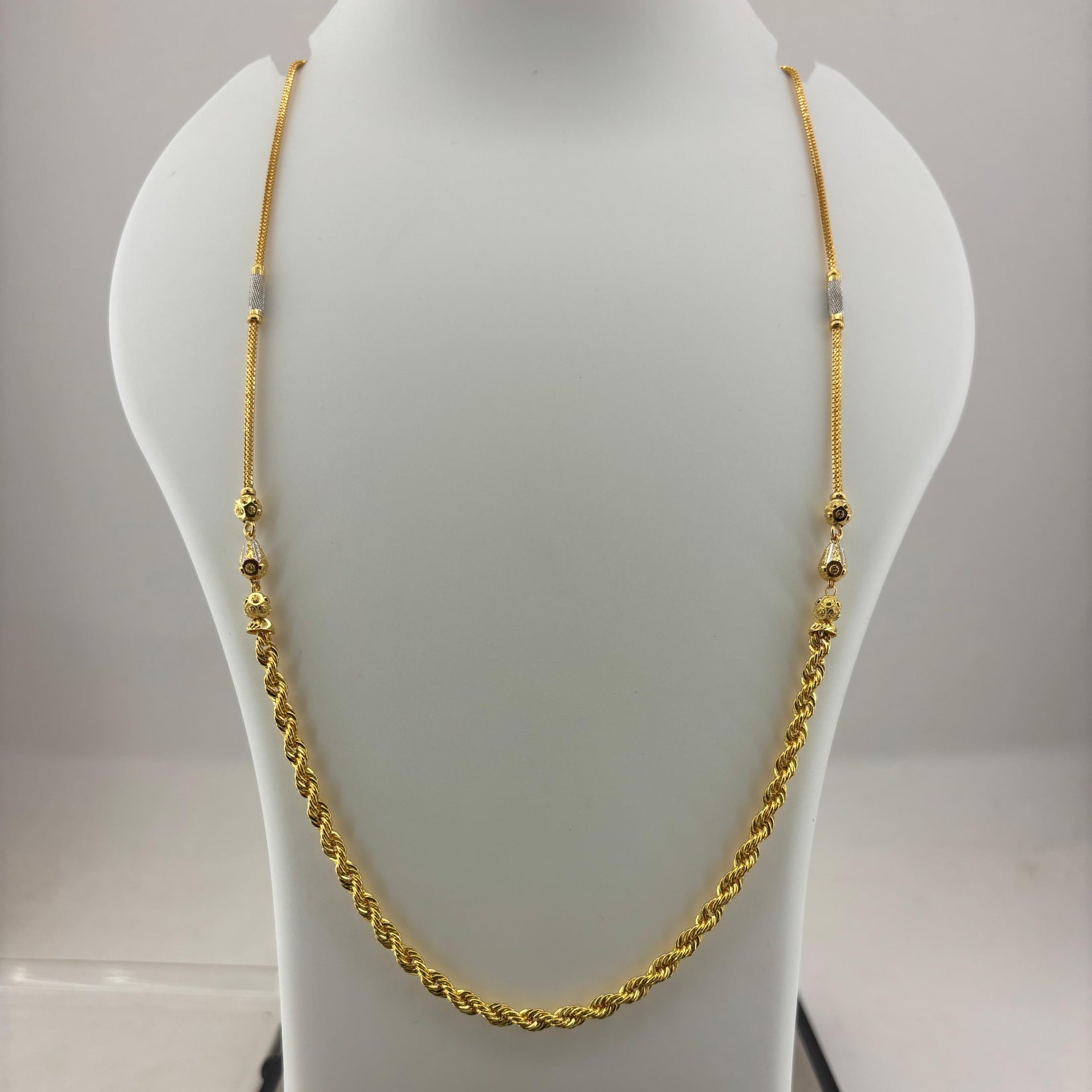 A gold chain necklace displayed on a white mannequin bust, with a digital scale visible underneath. The necklace has a twisted rope style with small bead accents.