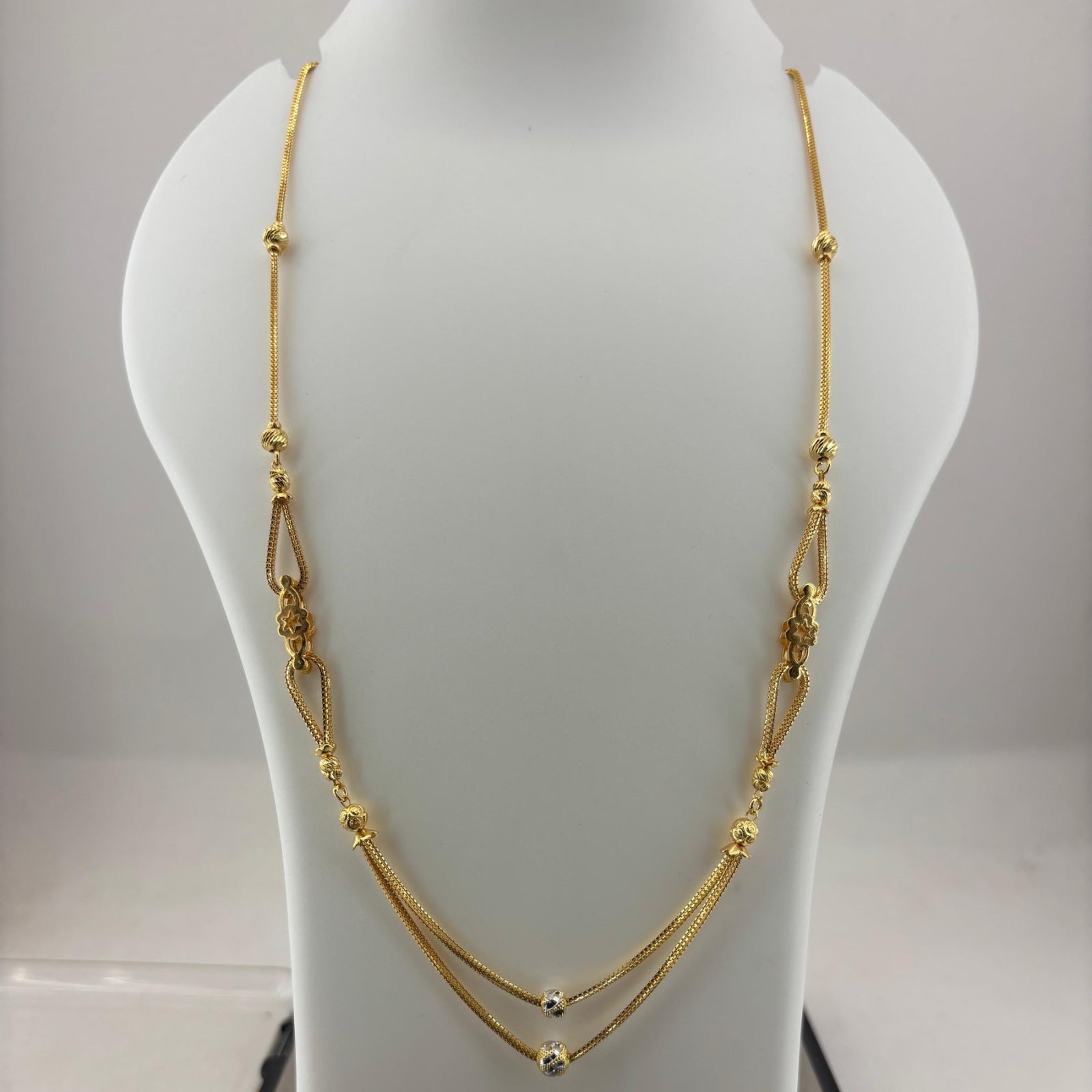 A gold-toned multi-strand necklace with textured bead details, displayed on a white jewelry bust stand with a digital scale underneath.