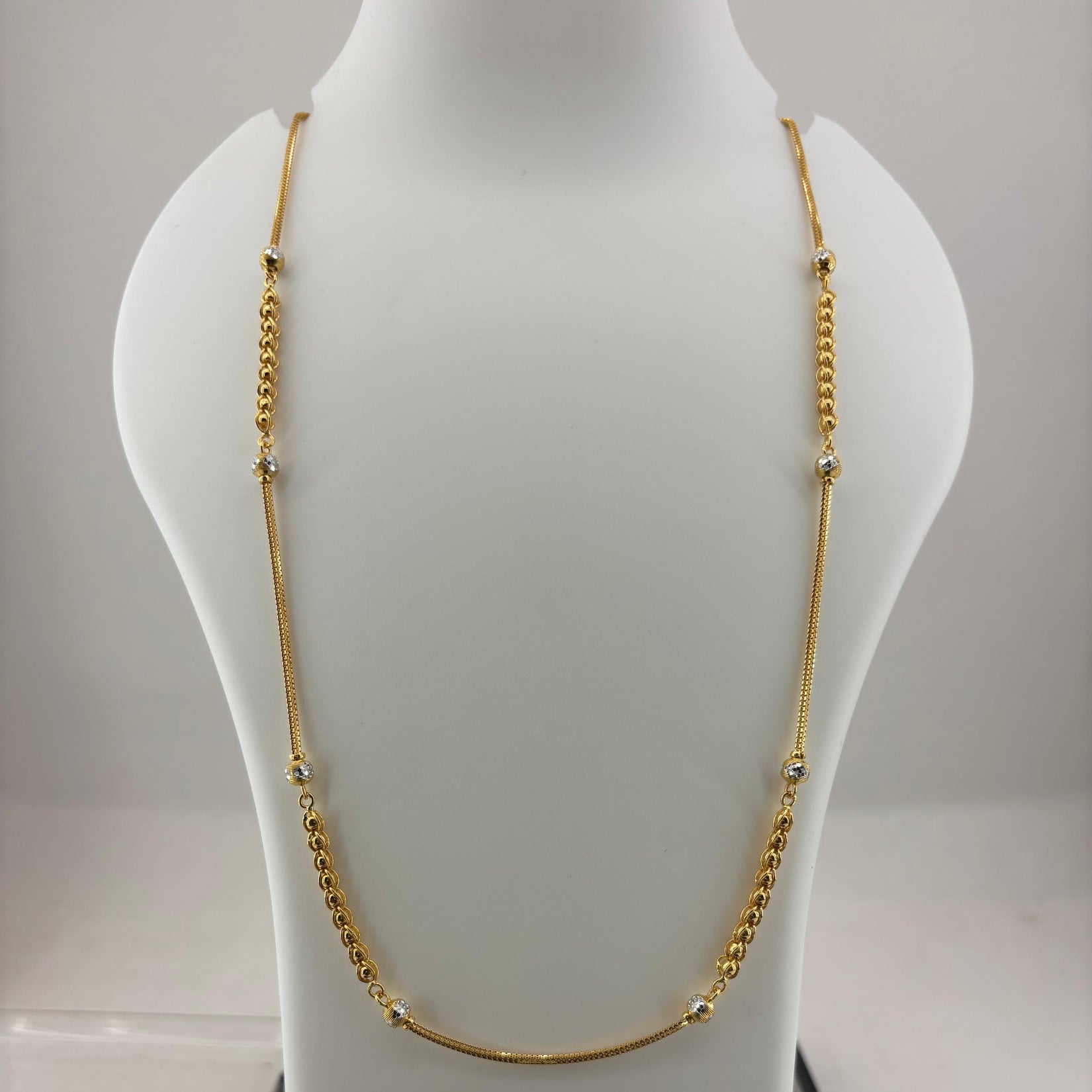 A gold chain necklace displayed on a white mannequin bust, with a digital scale visible underneath. The necklace features a smooth curved section at the bottom and small bead details spaced along the chain.