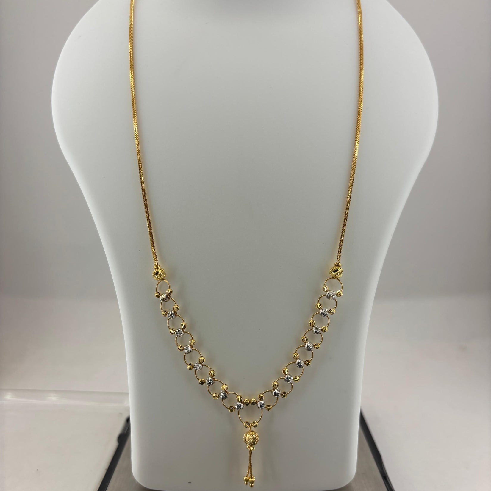 A gold necklace displayed on a white mannequin bust, with a digital scale visible underneath. The necklace features a delicate chain with a V-shaped arrangement of small gold links and a central pendant.