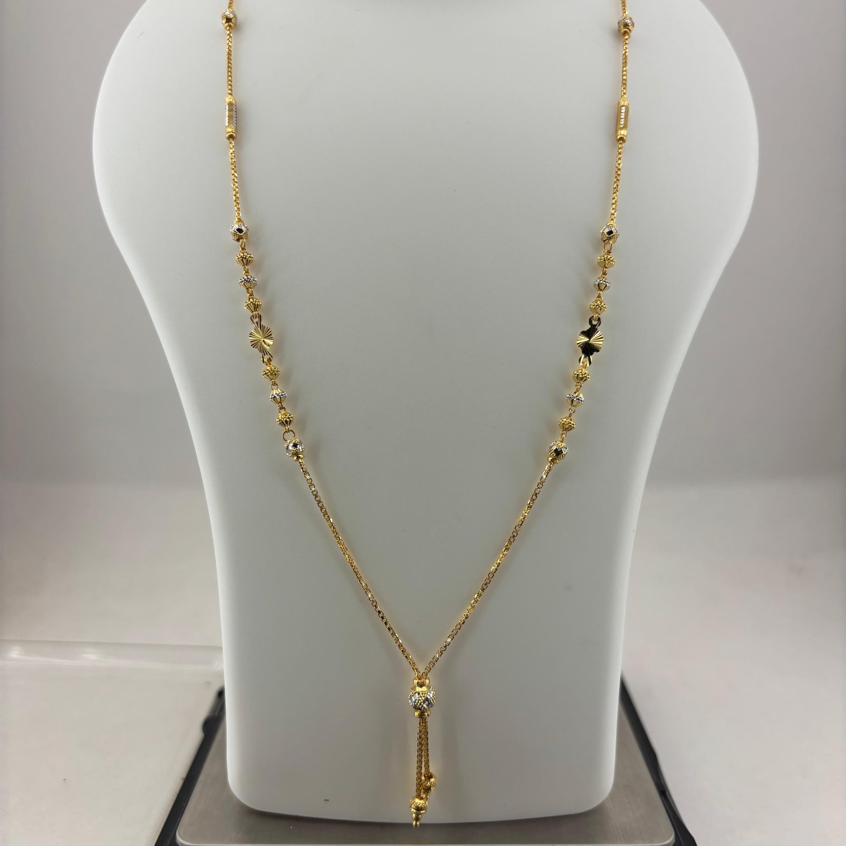 A gold necklace displayed on a white mannequin bust, with a digital scale visible underneath to show the weight. The necklace features a delicate chain with small bead details and a central pendant.