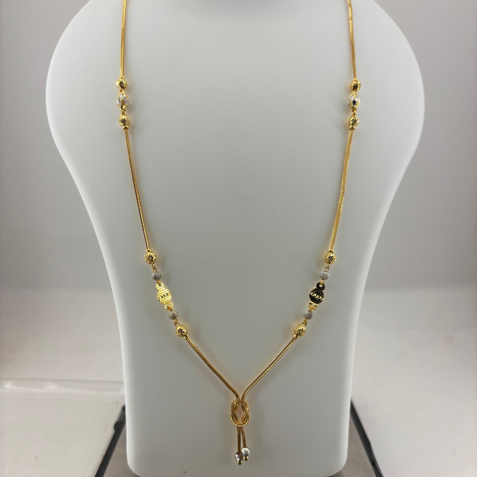 A gold necklace with small metallic beads and a key-shaped pendant, displayed on a white jewelry bust that sits on a digital scale.