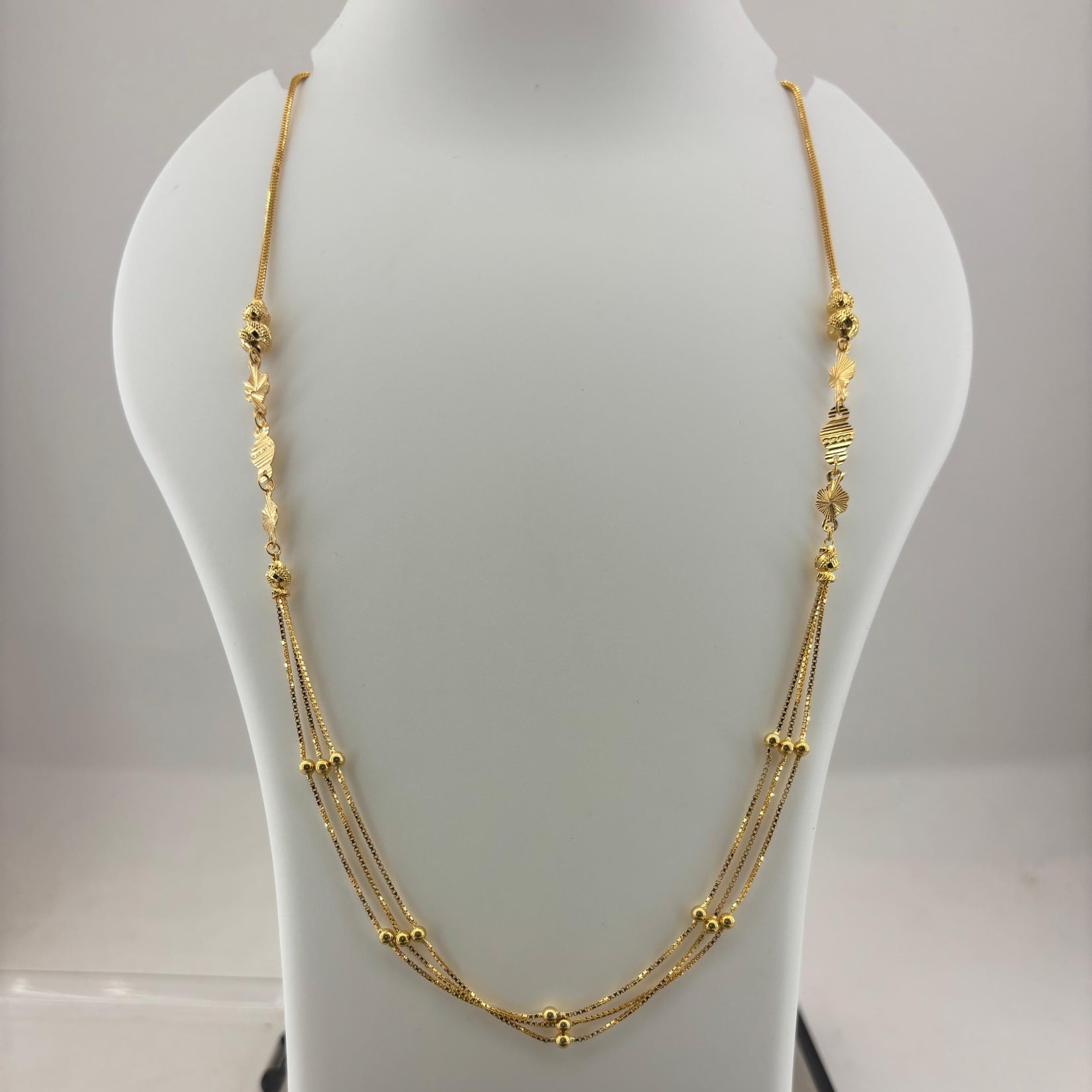 A gold multi-strand necklace displayed on a white mannequin bust, with a digital scale visible underneath. The necklace features multiple layers of chain with small gold bead and textured details.