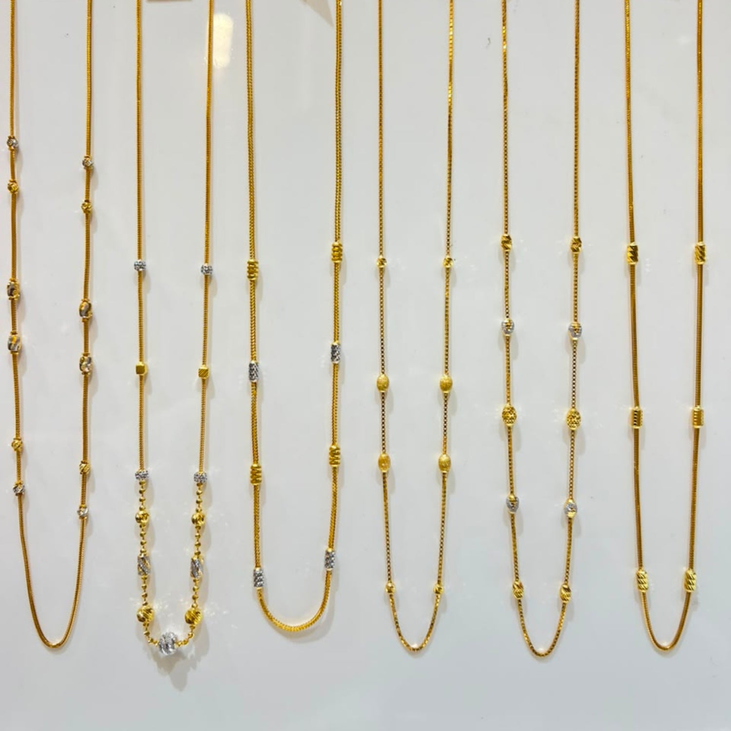 Six gold-toned chain necklaces with mixed metal bead and link details are displayed on a white jewelry stand, each with a small white price tag attached.