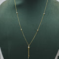A delicate gold necklace displayed on a dark green velvet jewelry bust. The necklace features a fine chain with small spaced gold beads and a central teardrop-shaped gold pendant from which a short chain dangles.
