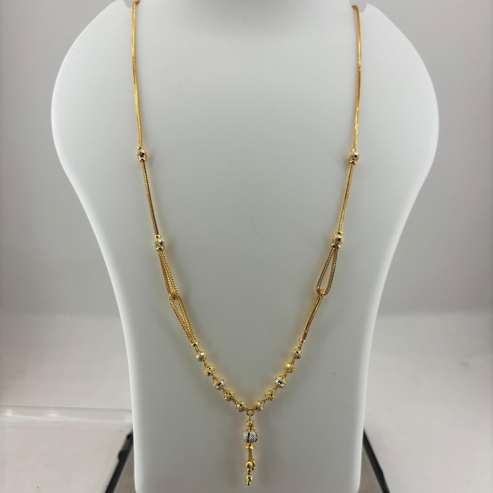 A gold necklace displayed on a white jewelry bust, with a digital scale visible underneath. The necklace features a delicate chain with small bead details and a central pendant with a clear stone.