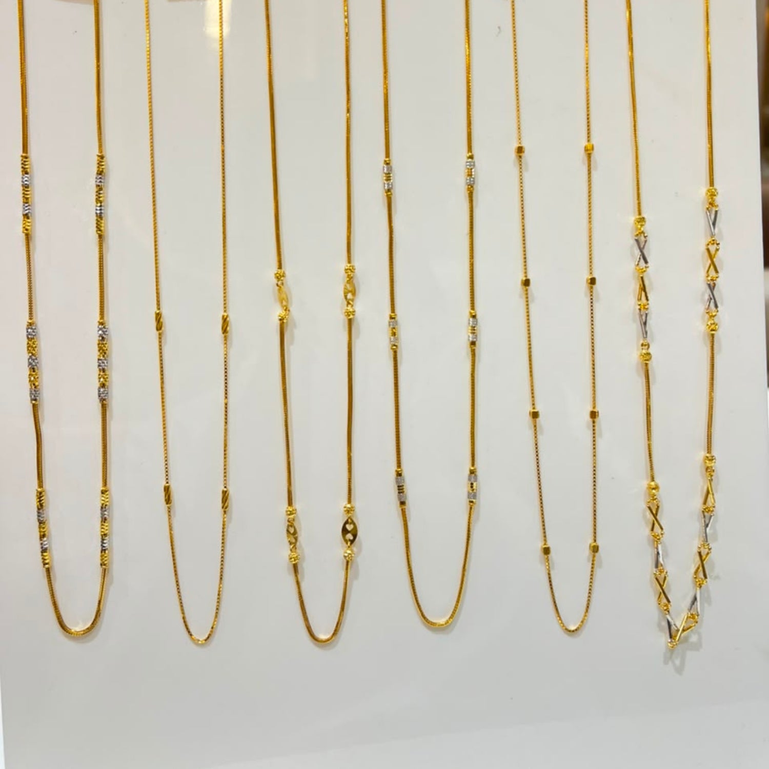Nine gold chains of varying lengths and styles displayed on a white stand, each with a small price tag attached.