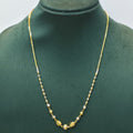 A delicate gold chain necklace with small alternating gold and white beads, featuring a central gold medallion, displayed on a dark green jewelry bust.