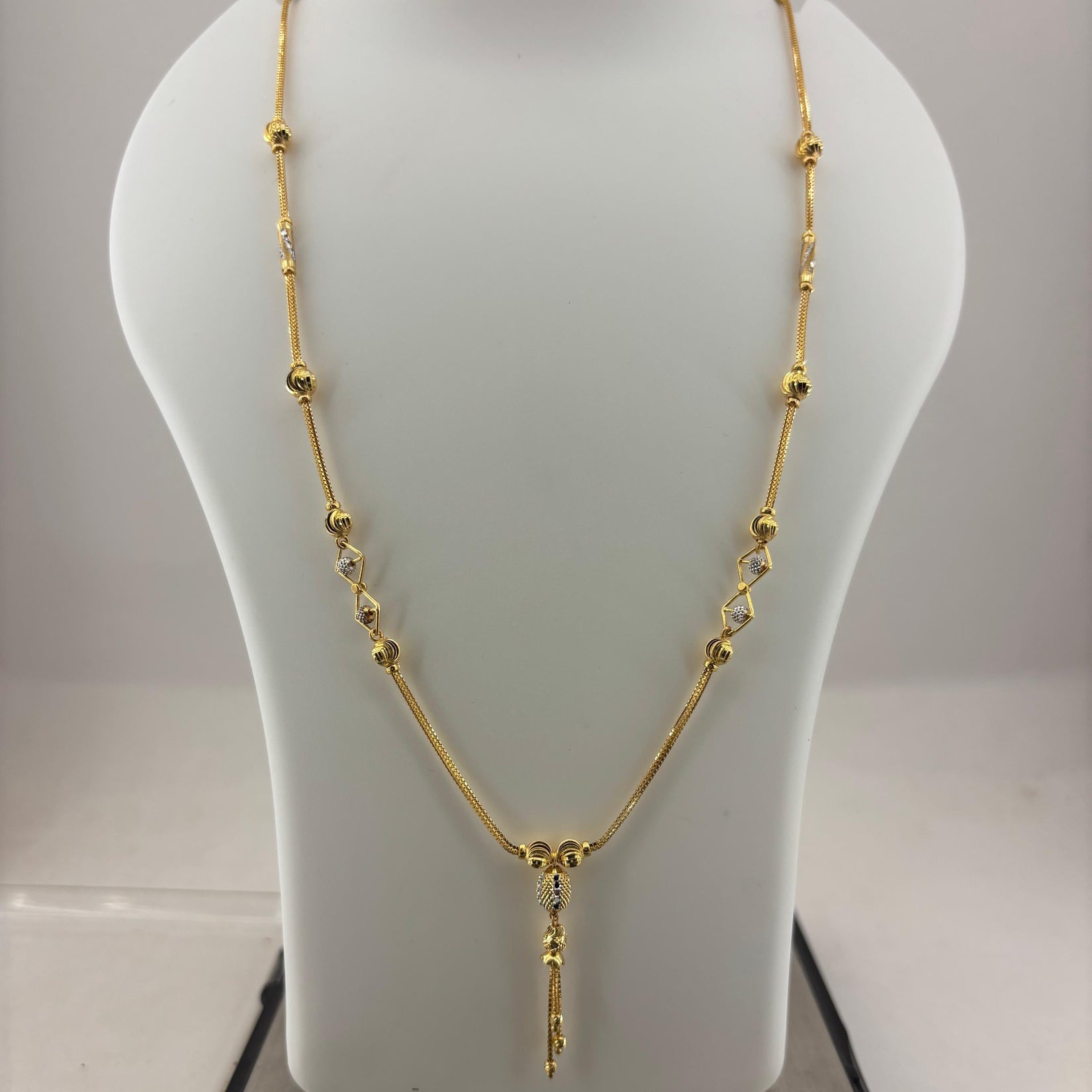 A gold necklace displayed on a white jewelry bust, with a digital scale visible underneath. The necklace features a delicate chain with small gold beads and a central pendant.