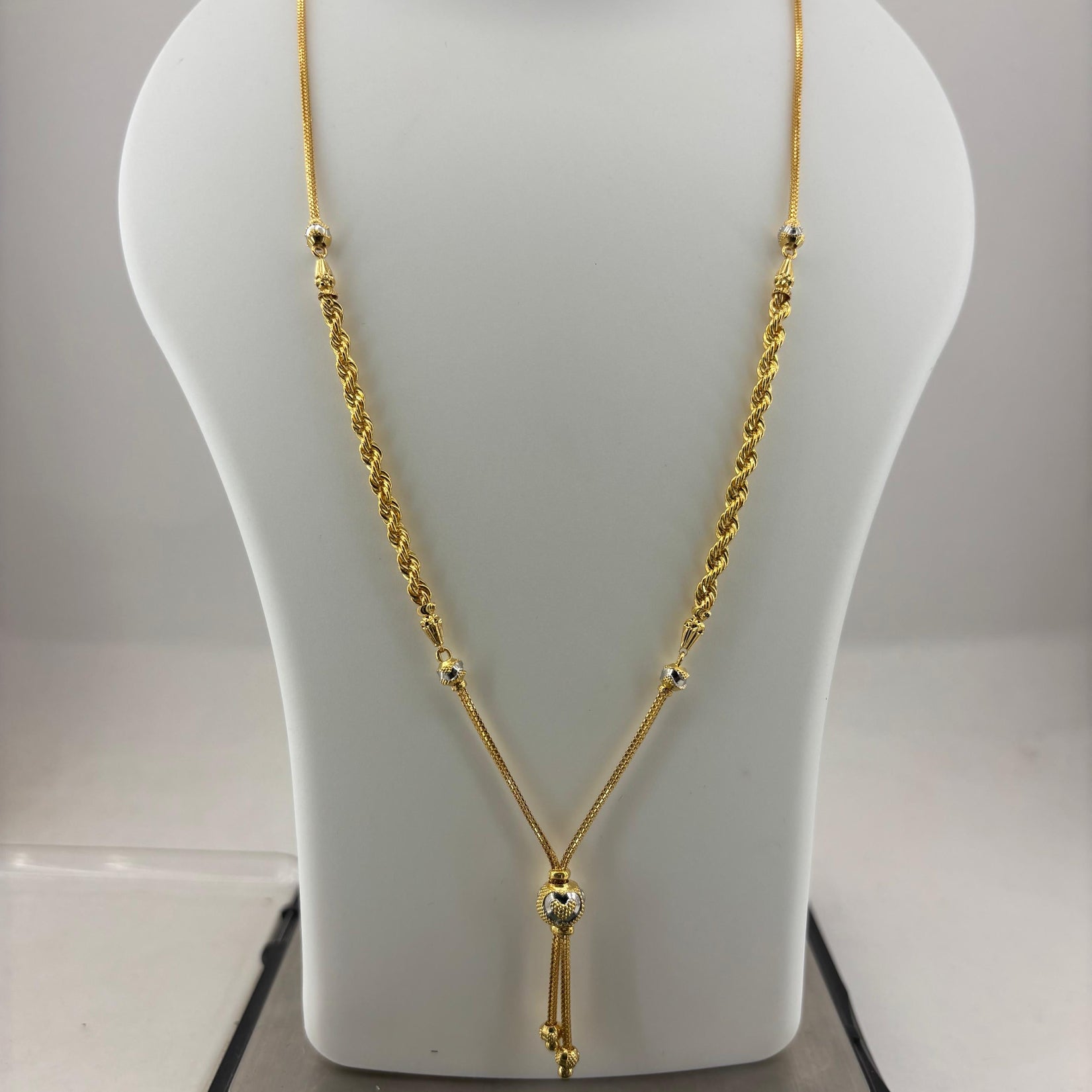 A gold necklace displayed on a white mannequin bust, with a digital scale visible underneath. The necklace features a twisted rope chain, small bead details, and a central pendant with hanging tassels.