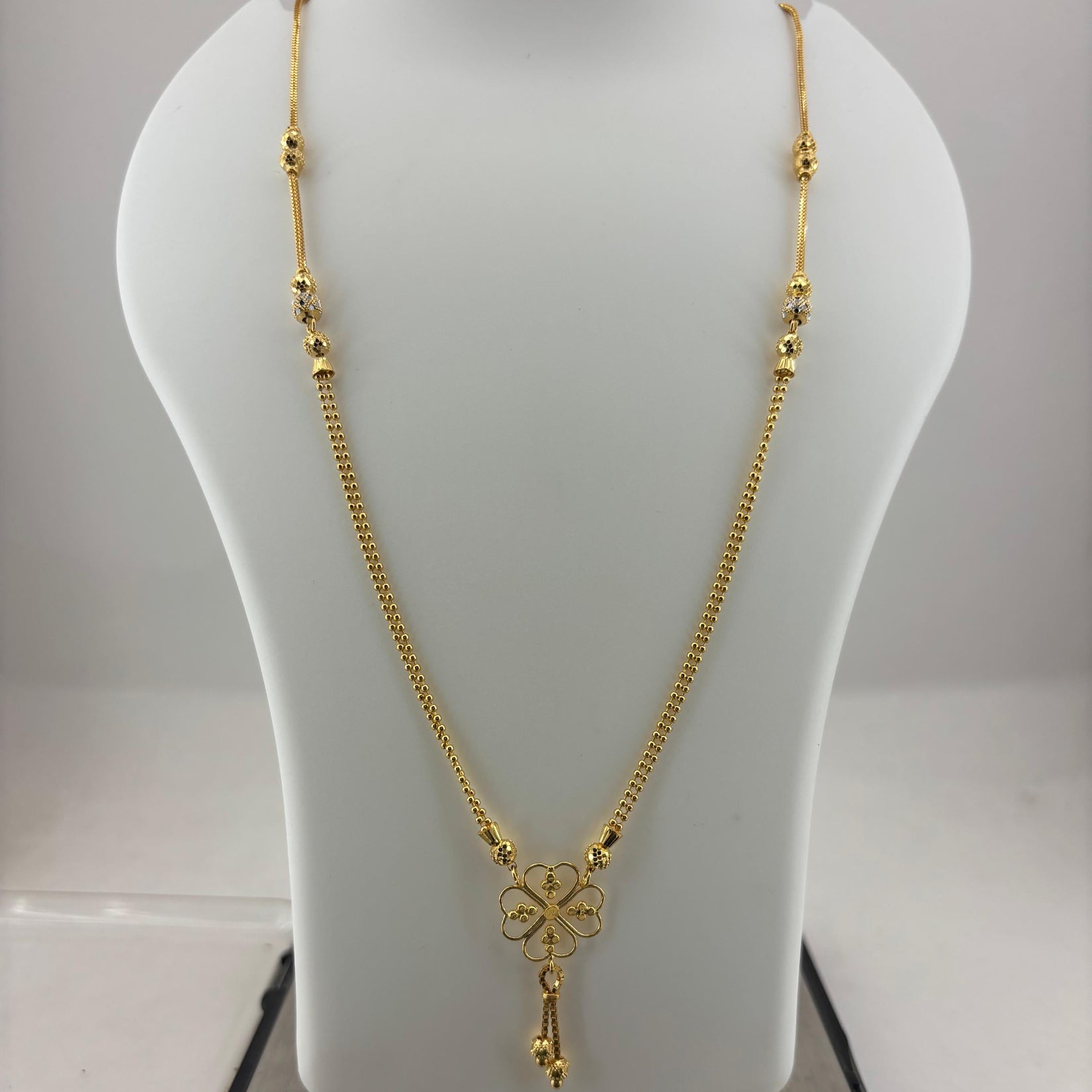 A gold necklace with a four-leaf clover pendant displayed on a white mannequin bust, with a digital scale visible underneath.