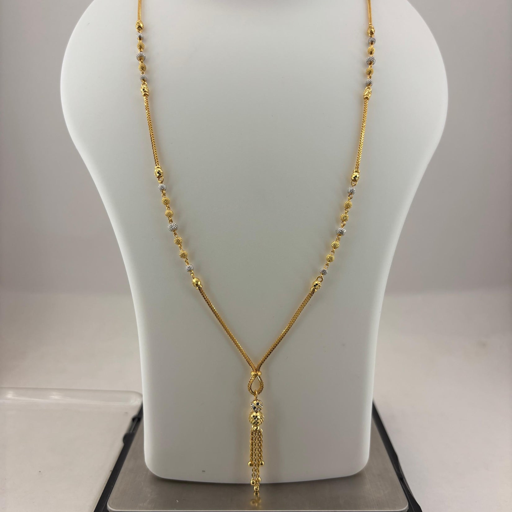 A gold necklace displayed on a white mannequin bust, with a digital scale visible underneath. The necklace features a delicate chain with small bead-like details and a textured pendant.