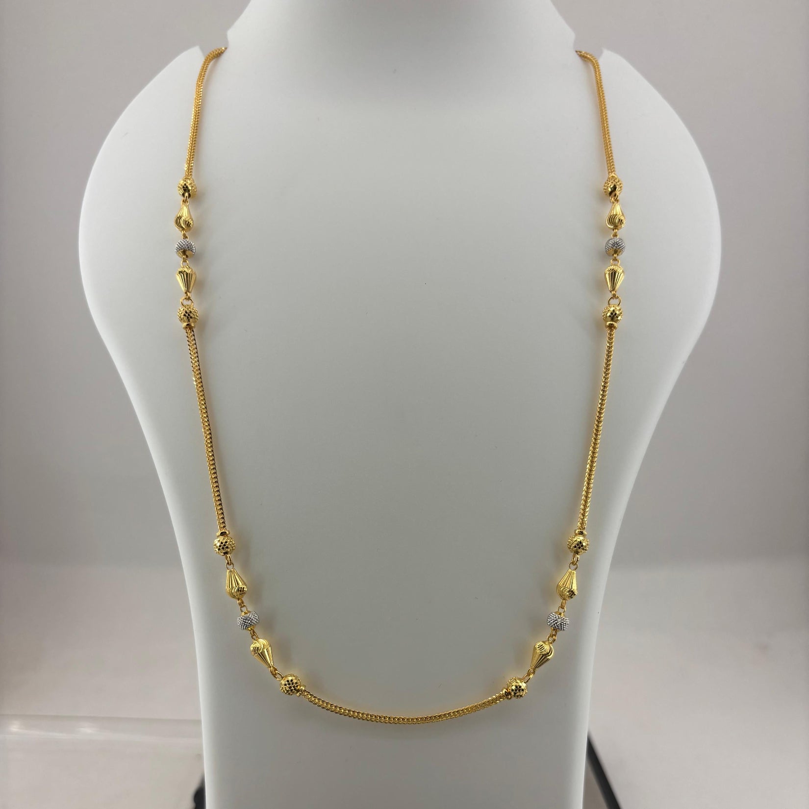 A gold chain necklace with small bead accents displayed on a white mannequin bust, with a digital scale visible underneath.