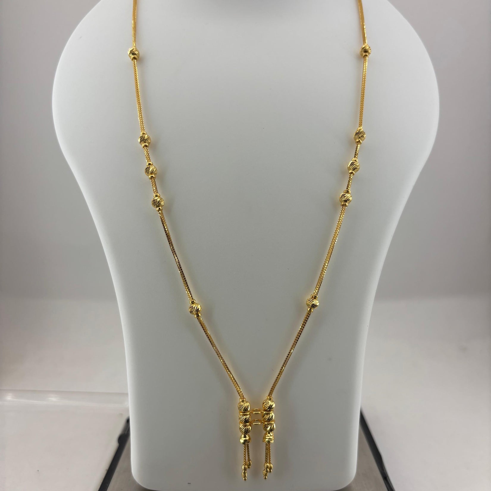 A gold necklace displayed on a white mannequin bust, with a digital scale visible underneath. The necklace features a delicate chain with small bead-like accents and a central pendant with three dangling chain ends.