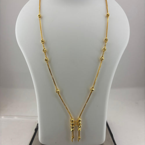A gold necklace displayed on a white mannequin bust, with a digital scale visible underneath. The necklace features a delicate chain with small bead-like accents and a central pendant with three dangling chain ends.