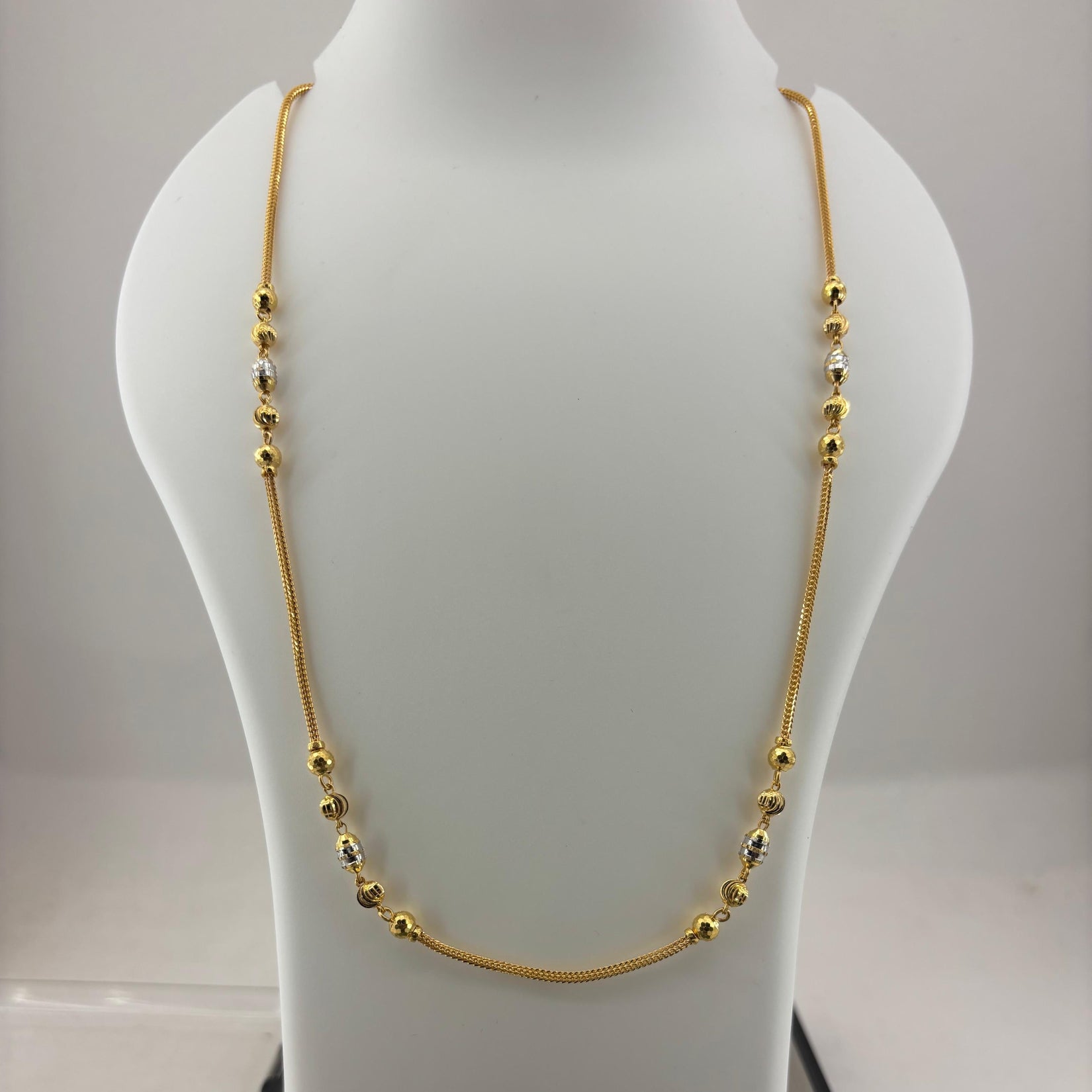 A gold chain necklace with small bead accents displayed on a white mannequin bust, with a digital scale visible underneath.