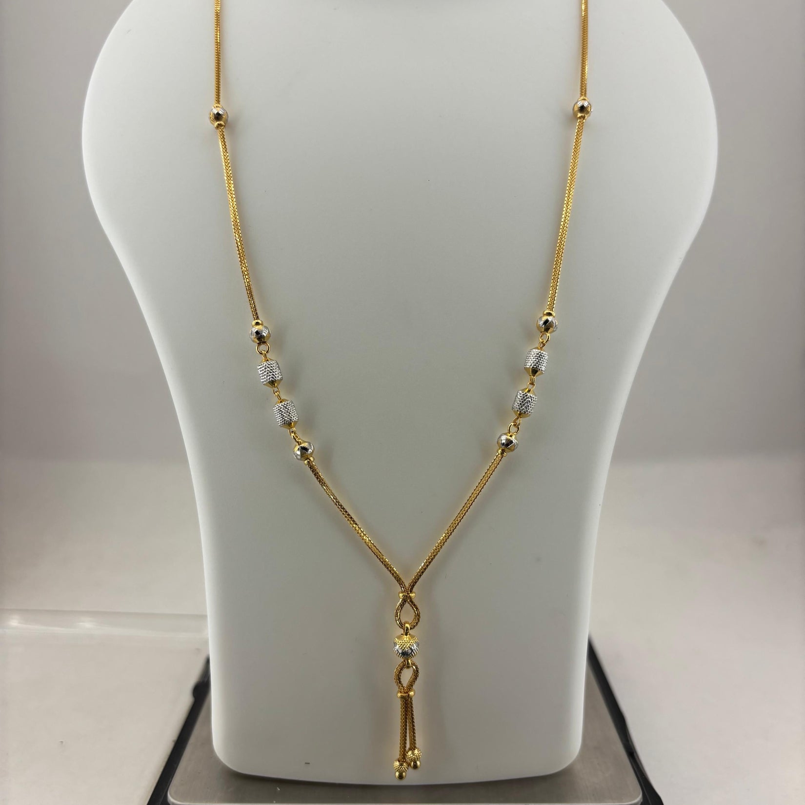 A gold chain necklace with small clear beads and a dangling gold tassel pendant, displayed on a white mannequin bust resting on a digital scale.