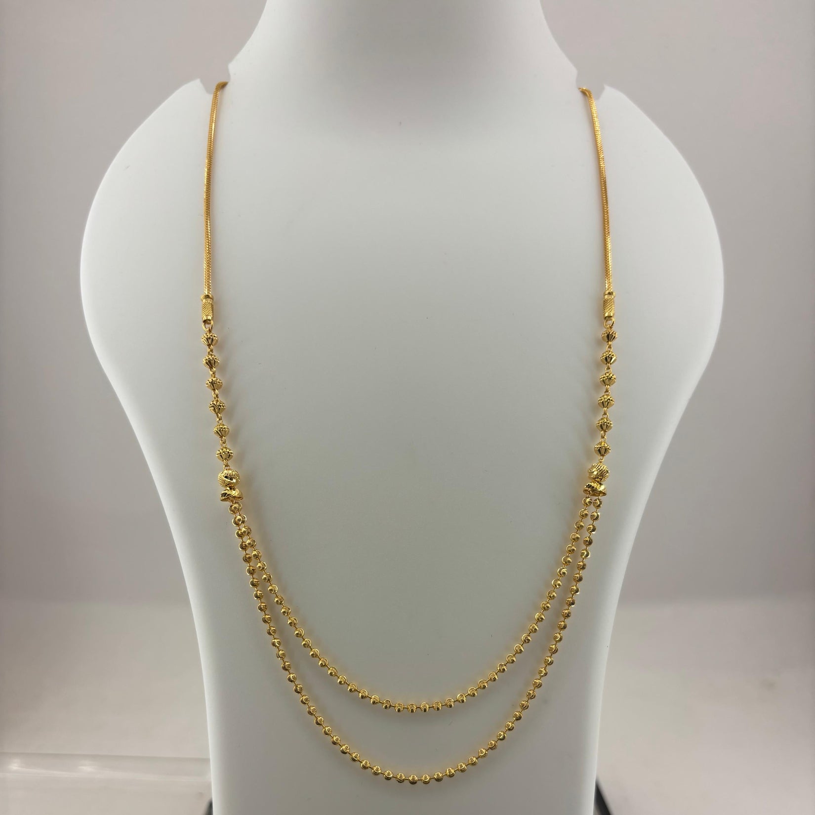 A double-strand gold chain necklace displayed on a white jewelry bust, with a digital scale visible underneath.