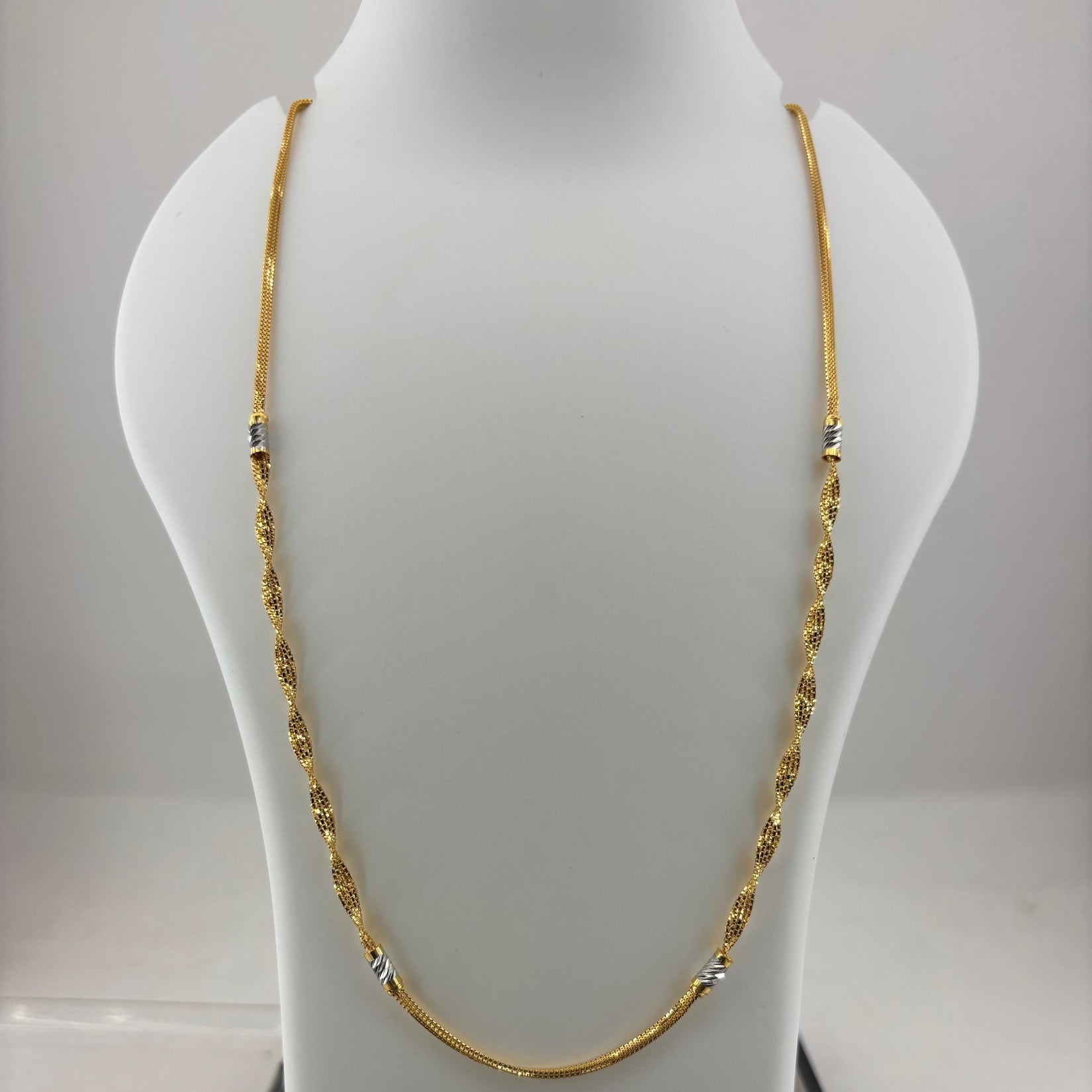 A gold chain necklace displayed on a white mannequin bust, with a digital scale visible underneath. The necklace has small silver-toned bead accents spaced along the chain.
