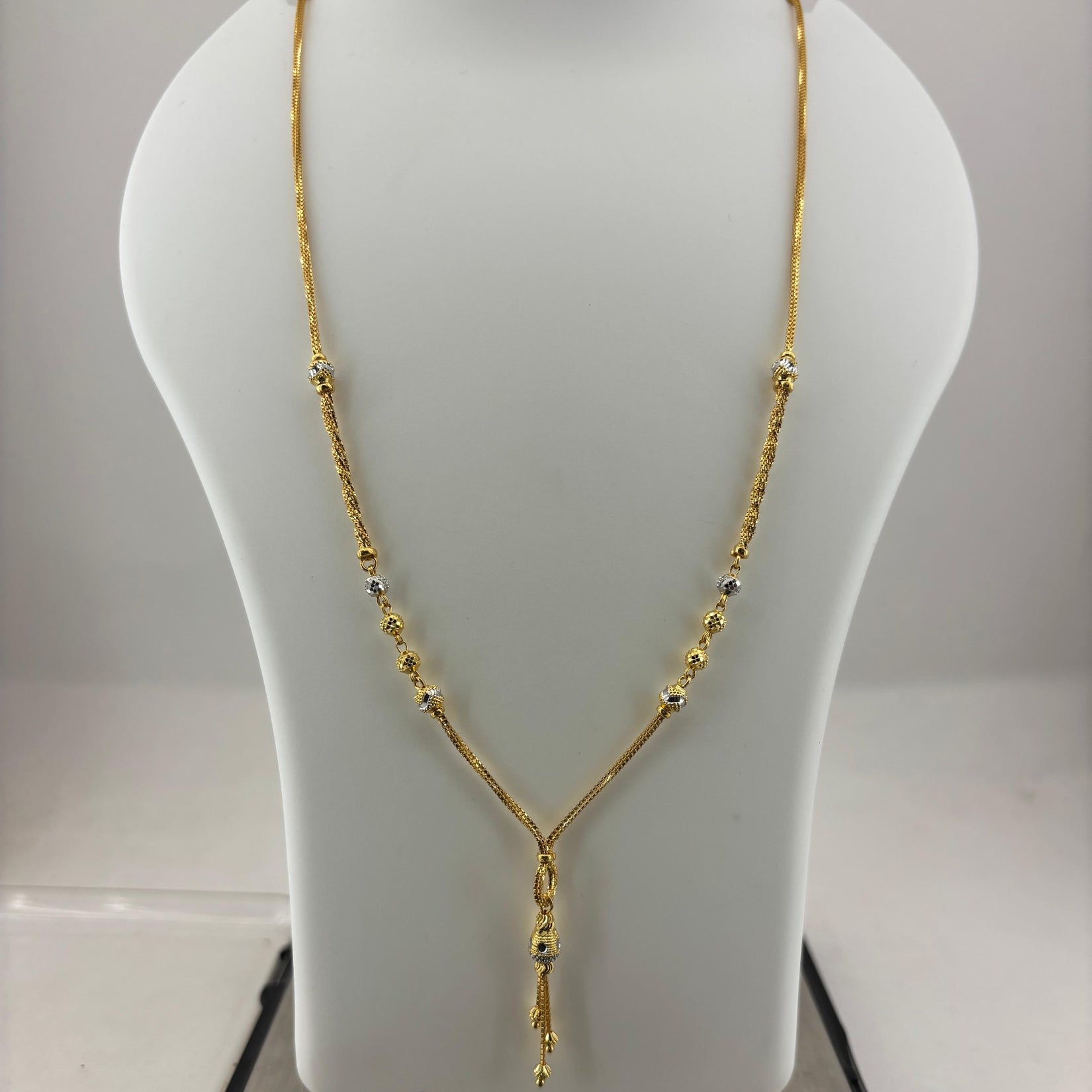 A gold necklace displayed on a white jewelry bust, with a digital scale visible underneath. The necklace has a Y-shaped design with small bead details along the chain.