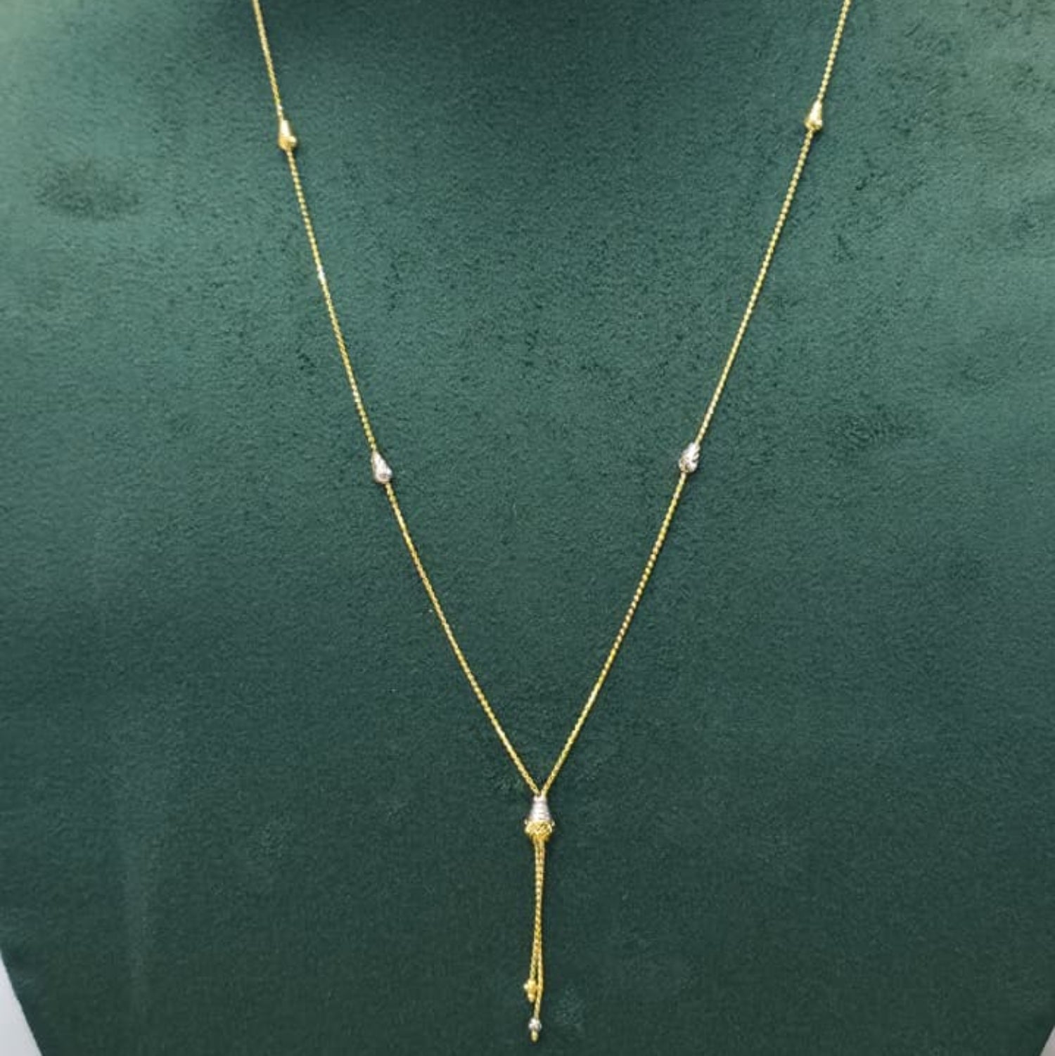 A delicate gold necklace with small bead details and a teardrop-shaped pendant, displayed on a dark green jewelry bust.