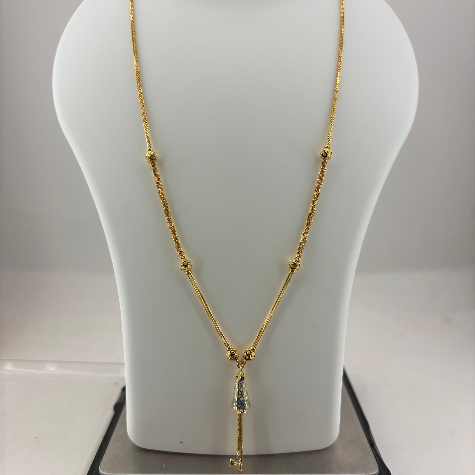 A gold necklace displayed on a white mannequin bust, with a digital scale underneath to show weight. The necklace features a delicate chain with small bead details and a central pendant with a blue-green stone.