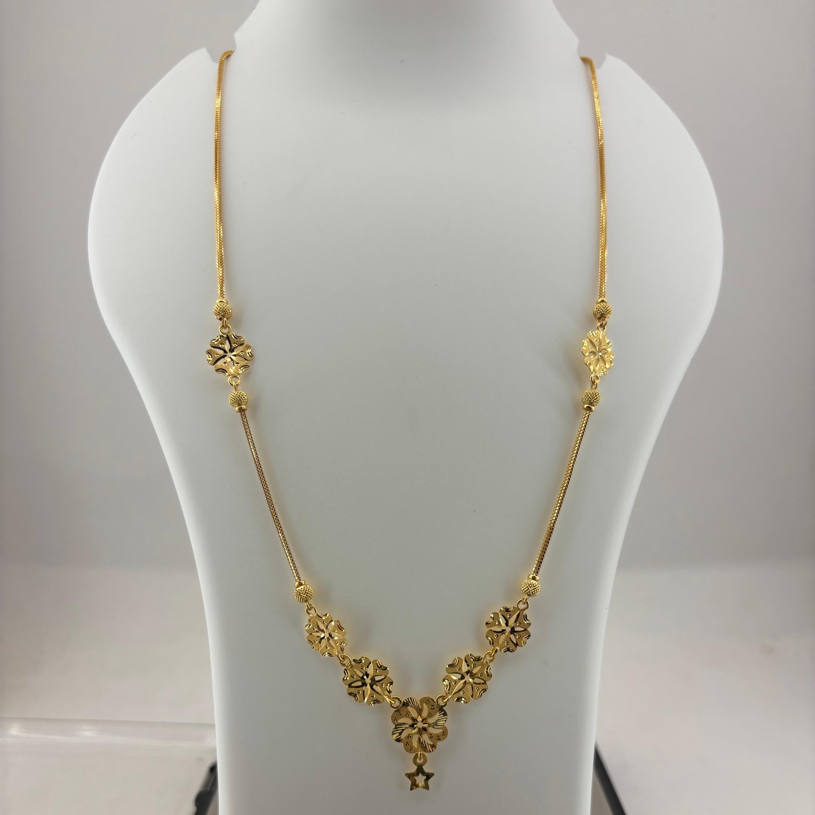 A gold necklace displayed on a white mannequin bust, with a digital scale visible underneath. The necklace features a delicate chain with five ornate gold floral clusters arranged in a V-shape at the center, culminating in a small star pendant.