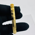A gold-toned bangle bracelet with geometric and floral engravings, held by a hand wearing a black glove against a plain light gray background.