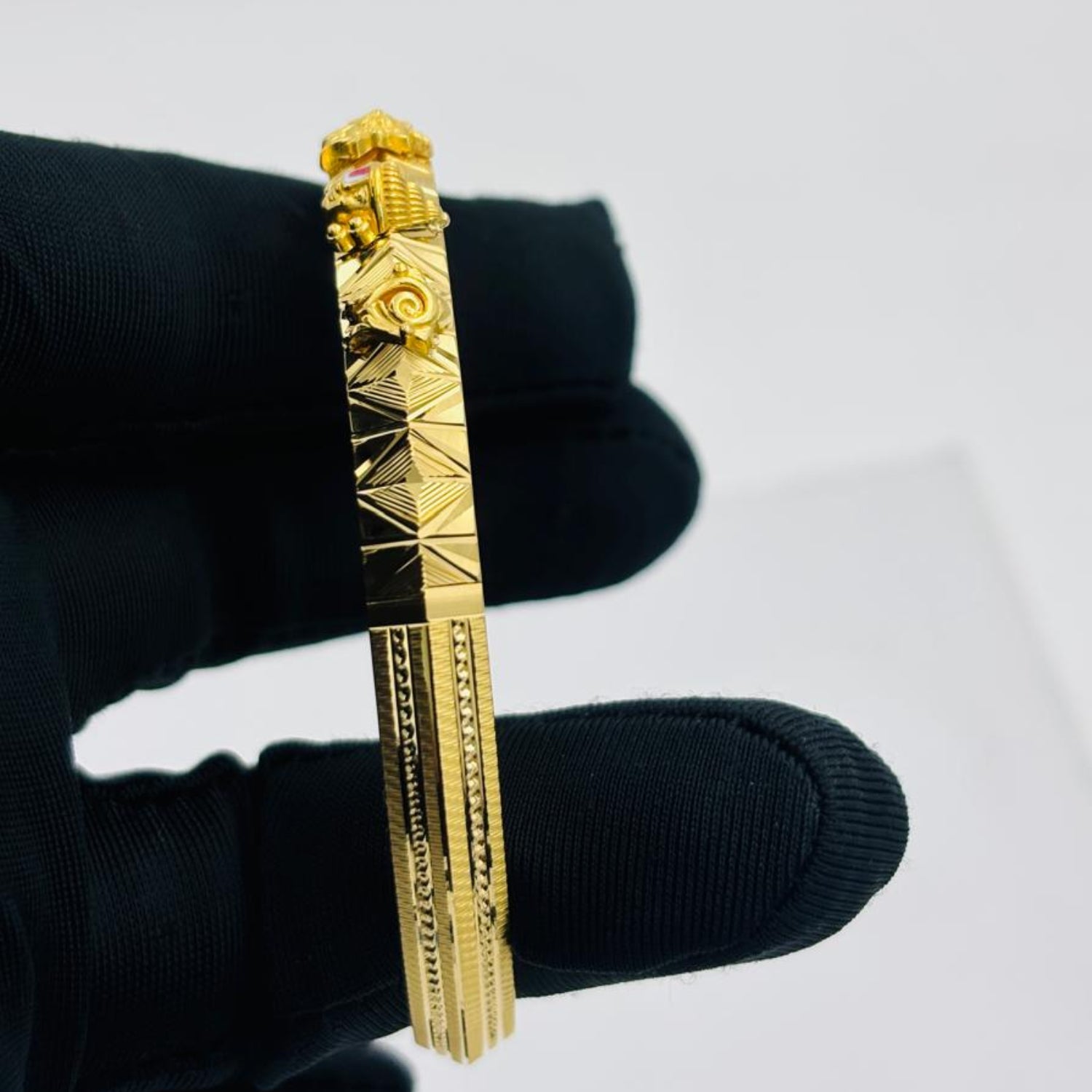 A gold-toned bangle bracelet with geometric and floral engravings, held by a hand wearing a black glove against a plain light gray background.
