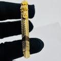A gold-toned rectangular bar pendant or brooch with ornate sculpted details and textured geometric sections, held between fingers wearing a black glove against a plain light gray background.
