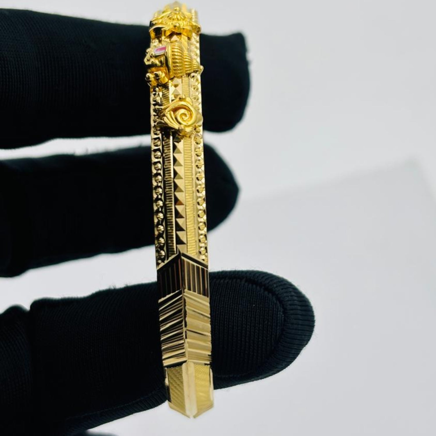 A gold-toned rectangular bar pendant or brooch with ornate sculpted details and textured geometric sections, held between fingers wearing a black glove against a plain light gray background.