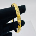 A gold-plated bangle bracelet with geometric patterns and small hanging decorative charms, held by a hand wearing a black glove against a light gray background.
