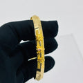 A gold-plated bangle bracelet with geometric and floral engravings, held by a hand wearing a black glove against a plain white background.