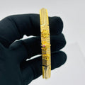A gold-plated bangle bracelet with intricate floral and geometric engravings, held by a hand wearing a black glove against a plain white background.