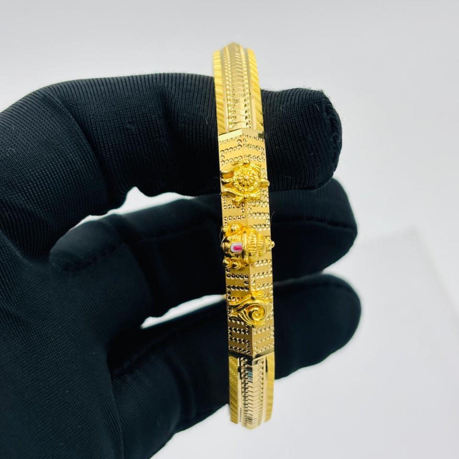 A gold-plated bangle bracelet with intricate floral and geometric engravings, held by a hand wearing a black glove against a plain white background.