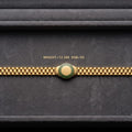 A gold-tone watch band with a central round green and gold emblem, displayed horizontally on a dark gray textured background. Small gold text above the band reads 'WEIGHT=12 GM SQ8-92'.
